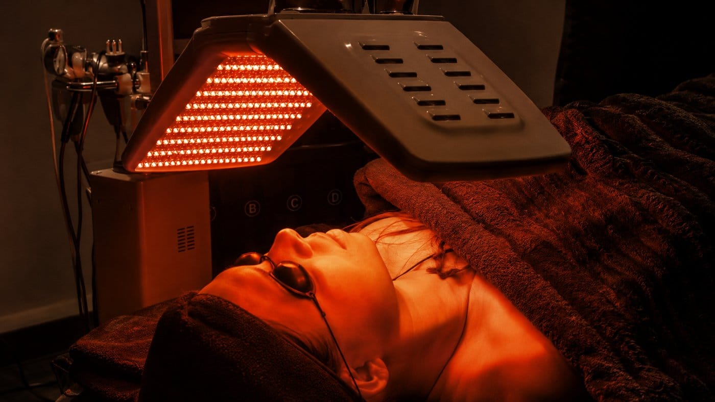 Client relaxing under a red LED light therapy panel during a facial treatment